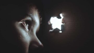 صورة لصبي يراقب من خلال ثقب في الحائط، تظهر عينيه بوضوح وتعكس شعور الفضول والدهشة. الضوء يتسلل من الثقب، مما يخلق تباينًا بين الظلام والنور.