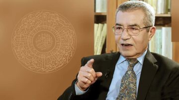 رجل مسن يرتدي بدلة رسمية وقميص بألوان هادئة، يتحدث بحماس ويشير بإصبعه. خلفية الصورة تحتوي على نمط هندسي عربي تقليدي.