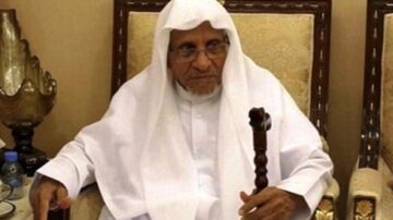 رجل مسن يرتدي الثوب والعمامة التقليدية، يجلس على كرسي مزخرف في غرفة ذات ديكور أنيق، ممسكًا بعصا، ويبدو عليه التعبير الجاد.