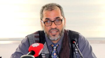 رجل يتحدث في ندوة، يرتدي سترة رمادية وقميص بنفسجي، ويضع نظارات. أمامه ميكروفون وميكروفون آخر باللون الأحمر. الخلفية بيضاء.