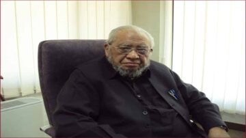 رجل مسن ذو لحية يجلس في مكتب، يرتدي زيًا أسود، ويبدو بملامح جدية. خلفه ستائر بيضاء تسمح بدخول الضوء الطبيعي، مما يضفي أجواء مهنية.