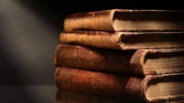 أربعة كتب قديمة متراكمة، تتميز بغلاف جلدي بني ومظهر تاريخي، مع حواف ممزقة وملمس ناعم، مضاءة بشكل جميل من جانب، تعكس التراث الأدبي والثقافي.