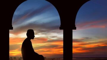 صورة ظلية لشخص جالس تحت قوسين مع غروب الشمس في الخلفية. السماء ملونة بتدرجات زاهية من البرتقالي والأزرق، مما يخلق جوًا هادئًا وروحيًا.