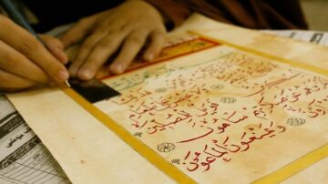 كتابة نص عربي بخط اليد على ورقة قديمة، مع التركيز على تفاصيل الحروف والزخارف. تظهر يد شخص يستخدم قلم على الصفحة، محاطة بأوراق صحفية.