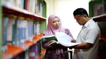 امرأتان ترتديان الحجاب، تقفان في مكتبة بين رفوف الكتب، وتتناقشان حول محتوى كتاب مفتوح، مع التركيز على التعلم والمعرفة.