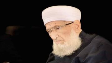 شيخ مسن ذو لحية بيضاء يرتدي عمامة بيضاء ونظارات، يظهر بتعبير هادئ ومتفكر في بيئة داكنة.