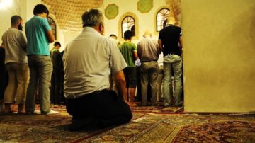 رجل جالس على ركبتيه في مسجد، محاط بمجموعة من المصلين الذين يقفون في صفوف أثناء الصلاة. تظهر تفاصيل السجاد التقليدي وزخارف الجدران في الخلفية، مما يبرز الأجواء الروحية للمكان.