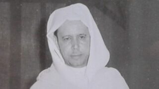 صورة تاريخية لرجل يرتدي جلباب أبيض وعمامة، يظهر بملامح جادة، في خلفية غير واضحة. تعكس الصورة الطابع الثقافي والزي التقليدي.
