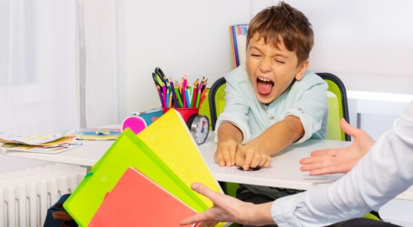 طفل صغير يجلس على مكتب ويظهر تعبيرًا عن الإحباط أو الغضب أثناء محاولته سحب ملفات ملونة من يد شخص بالغ. في الخلفية، أدوات مكتبية وأدوات فنية مرتبة.