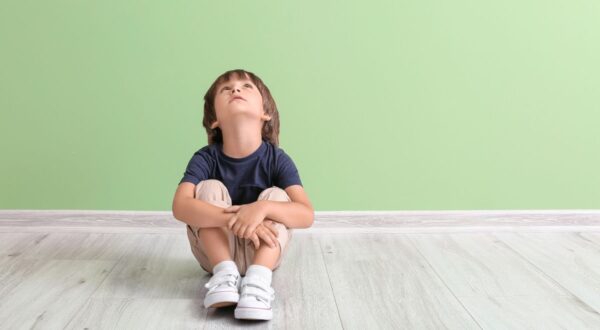 طفل صغير يجلس على الأرض، ينظر إلى الأعلى، مع تعبير فضولي، في غرفة ذات جدار أخضر فاتح. يرتدي قميصًا داكنًا وشورتات فاتحة، مع حذاء رياضي أبيض. الأرضية خشبية فاتحة.