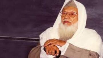 رجل مسن ذو لحية بيضاء يرتدي عمامة وقميصًا تقليديًا، يجلس مع عصا في يده، يظهر عليه وقار وحكمة.