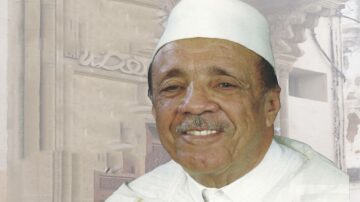 رجل مسن مبتسم يرتدي غطاء رأس أبيض، يظهر خلفه معلم تاريخي. تعكس الصورة التراث والثقافة، مما يبرز الهوية المحلية.