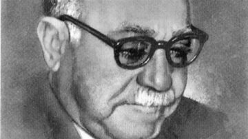 صورة توضح رجل مسن يرتدي نظارات داكنة، مع لحية وشعر رمادي. يركز الرجل على شيء أمامه، مظهره يعكس الحكمة والعمق. الخلفية بسيطة وغير مزخرفة، مما يبرز ملامحه.