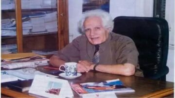 رجل مسن يجلس على مكتب في مكتب، محاط بمجلات وملفات. يحمل فنجان قهوة ويبتسم. خلفية الصورة تحتوي على خزائن مليئة بالكتب والمستندات.