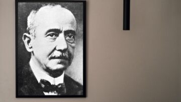 صورة قريبة لرجل مسن ذو شارب، يرتدي بدلة رسمية وربطة عنق. الصورة بالأبيض والأسود مؤطرة ومعلقة على حائط بلون محايد.