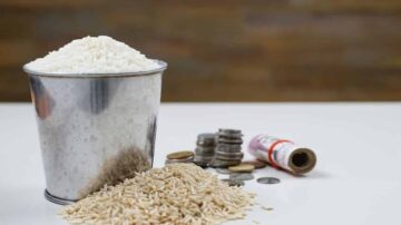 دلو معدني مليء بالأرز الأبيض، مع أرز بني متناثر بجانبه. ترتب بعض العملات المعدنية وبساطة لفافة نقدية بالقرب من الدلو. خلفية غير واضحة تضيف إلى التركيز على العناصر.