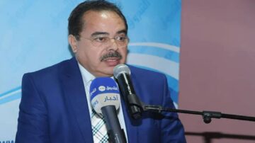 رجل يرتدي بدلة زرقاء يتحدث إلى ميكروفون خلال حدث رسمي، مع وجود خلفية تحمل شعار مؤسسة أو منظمة.