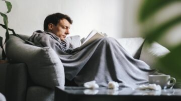 رجل يجلس على أريكة مريحة، مغطى ببطانية رمادية، يقرأ كتابًا بتركيز. في الخلفية، توجد نباتات منزلية وكوب على الطاولة. تعكس الصورة أجواء من الاسترخاء والهدوء.