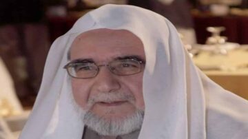 رجل مسن يرتدي غطاء رأس أبيض وعباءة، يظهر في بيئة داخلية مع خلفية غير واضحة. يعبر عن الحكمة والاحترام، مع نظارات وقصة شعر بيضاء.