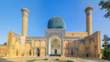 واجهة ضريح تيمورلنك في سمرقند، أوزبكستان، تظهر قبة زرقاء مزخرفة، وبرجين مميزين مزينين بنقوش ملونة، مع سماء زرقاء صافية.