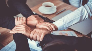 أيدي مجموعة من الأشخاص تتداخل فوق طاولة عمل، مع وجود فنجان من القهوة وأجهزة كمبيوتر محمولة في الخلفية، تعبر عن التعاون والعمل الجماعي في بيئة مهنية.