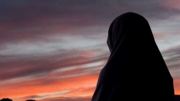 امرأة ترتدي حجابًا أسودًا تقف أمام غروب شمس ملون، مع سماء مفعمة باللون الأحمر والبرتقالي، مما يخلق مشهدًا هادئًا وجميلًا.