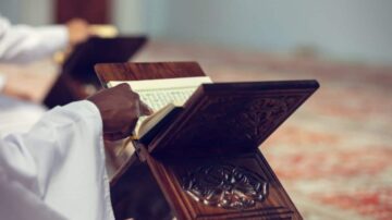 رجل يرتدي ثوبًا تقليديًا يقرأ القرآن الكريم من مصحف موضوع على حامل خشبي مزخرف. تظهر خلفية هادئة مع سجادة الصلاة.