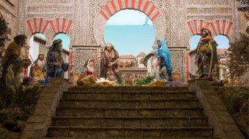 مشهد لمشهد ميلاد تقليدي يظهر تماثيل لعائلة يسوع المقدسة، مريم ويوسف، ومجموعة من الشخصيات المحيطة بهم. الخلفية تتضمن تفاصيل معمارية مزخرفة، مع قوس كبير مزين. الألوان دافئة، وتظهر عناصر طبيعية مثل النباتات.