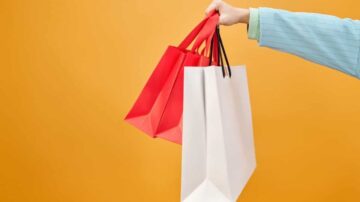 يد تحمل ثلاثة أكياس تسوق ملونة، اثنان أحمر وواحد أبيض، أمام خلفية برتقالية زاهية. أكياس التسوق تعكس أسلوب الحياة العصري والتسوق.