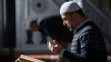 رجل يرتدي قميصًا داكنًا وقبعة بيضاء، يتلو القرآن في مسجد، يظهر التركيز والتأمل. في الخلفية، شخص آخر يتجه نحو السجود. الضوء الطبيعي يسلط على المشهد، مما يخلق جوًا روحيًا.