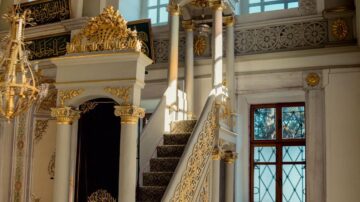 منظر داخلي لمسجد يظهر درجًا مزخرفًا باللونين الذهبي والأبيض يقود إلى منصة مرتفعة. تتزين الجدران بنقوش دقيقة وتفاصيل معمارية فاخرة، مع نوافذ تسمح بدخول الضوء الطبيعي.