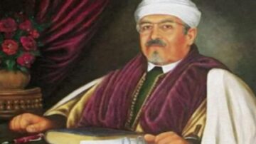 صورة لرجل يرتدي عمامة بيضاء وعباءة تقليدية، يجلس في غرفة مزينة بالستائر الحمراء وزهور الورد. يحمل كتابًا على الطاولة أمامه، ويظهر تعبيره الجاد والثقة.