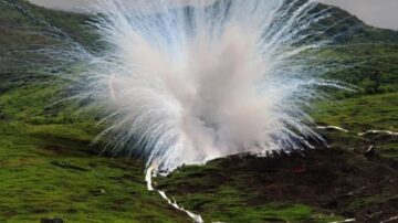 انفجار كبير يحدث في منطقة جبلية، حيث تتصاعد سحب من الدخان والغبار الأبيض من قمة التل الأخضر. تشكل المشهد تضاريس طبيعية رائعة، مع وجود تأثيرات بصرية مثيرة.