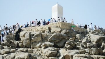 جبل الرحمة في عرفة، يظهر مجموعة من الحجاج يرتدون ملابس الإحرام، يتجمعون حول النصب التذكاري الأبيض على قمة الصخور. المنظر يعكس أجواء الحج والروحانية، مع سماء زرقاء وصخور كبيرة.