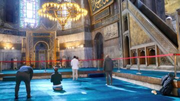 داخل مسجد تاريخي، زوار يتجولون على سجادة زرقاء مع وجود حواجز حمراء. في الخلفية، يُظهر الضوء من النوافذ الملونة تفاصيل معمارية مدهشة وسقف مزخرف.