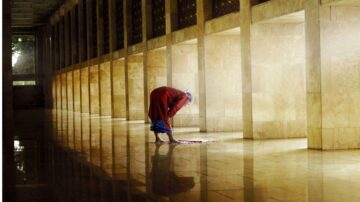 امرأة تصلي في ممر مضاء بأشعة الشمس، تعكس الأضواء على الأرض اللامعة. تظهر تفاصيل العمارة الحديثة مع جدران حجرية.