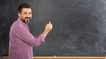 مدرس يبتسم وهو يحمل طبشورة في يده، يقف أمام سبورة سوداء فارغة، يرتدي قميصاً مخططاً.