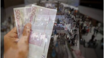 يد تظهر ورقتين نقديتين من فئة 500 يورو، مع خلفية غير واضحة لمشهد من سوق أو معرض مليء بالناس والأنشطة.