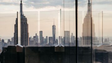 منظر لمدينة نيويورك يظهر ناطحات السحاب بما في ذلك مبنى إمباير ستيت وبرج الحرية، مع انعكاسات زجاجية تضيف عمقًا للمشهد. السماء ملبدة بالغيوم، مما يضفي جوًا دراميًا على الصورة.