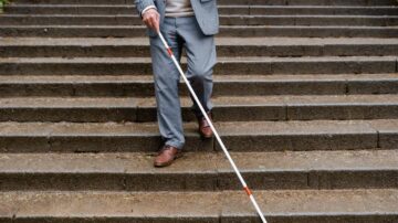 رجل يرتدي بدلة رمادية يسير على درج حجري باستخدام عصا بيضاء. يظهر التركيز والدقة في خطواته، مما يعكس أهمية أدوات التنقل للأشخاص ذوي الإعاقة البصرية.