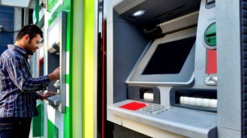 رجل يستخدم ماكينة صراف آلي في موقع حضري، مع وجود ماكينة أخرى قريبة. تظهر تفاصيل الشاشة وأزرار الماكينة بوضوح.