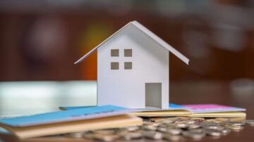 منزل ورقي صغير مع نوافذ، محاط بعملات معدنية ودفاتر ملونة. الصورة تعبر عن مفهوم الاستثمار العقاري والادخار.