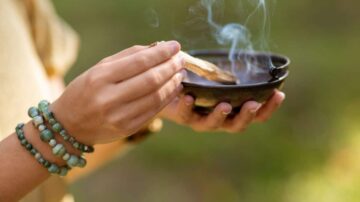 امرأة تحمل وعاء صغير يحتوي على بخور يتصاعد منه الدخان، ترتدي أساور ملونة على معصمها. الخلفية طبيعية وغير واضحة.