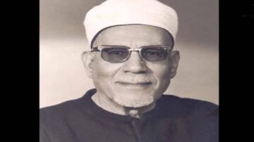 صورة لرجل مسن يرتدي عمامة بيضاء ونظارات شمسية، مبتسمًا. يظهر في الخلفية تدرجات لونية فاتحة، مما يبرز ملامحه. تعكس الصورة التراث الثقافي والديني.