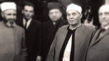 صورة تاريخية لأربعة رجال يرتدون ملابس تقليدية ورسمية، مع تركيز على رجل في المقدمة يرتدي جبة بيضاء وقبعة. الخلفية غير واضحة، مما يبرز الشخص في المقدمة.