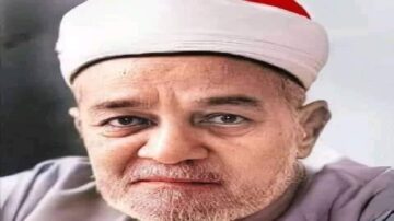 صورة لرجل مسن يرتدي عمامة بيضاء وقبعة حمراء، يظهر تعابير وجهه الجادة مع لحية رمادية. الخلفية بسيطة وغير مشغولة، مما يبرز ملامح وجهه.