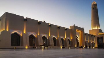 مبنى معماري معاصر مضاء بأضواء دافئة، يظهر تفاصيل النوافذ القوسية والمئذنة العالية. الأجواء هادئة مع سماء زرقاء فاتحة، تعكس جمال العمارة الإسلامية.