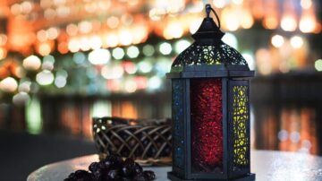 فوانيس ملونة مع خلفية مضيئة، تظهر فانوس أسود مزخرف مع ضوء أحمر داخله، بجانبه سلة تحتوي على فواكه. الأضواء الخلفية تضفي جوًا دافئًا ومناسبًا للأجواء الاحتفالية.