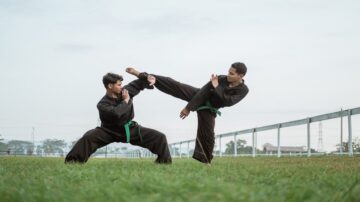 ممارسو فنون القتال يرتدون زيًا أسود مع أحزمة خضراء يتبادلون الحركات في ملعب أخضر. أحدهم ينفذ ركلة عالية بينما الآخر يتخذ وضعية دفاعية. السماء صافية والأفق يظهر خطوطًا كهربائية ومبانٍ في الخلفية.