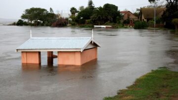 منزل جزئي مغمور بالمياه بسبب الفيضانات، مع الأشجار والمنازل في الخلفية. تعكس المياه السماء الرمادية، وتظهر أثار الأمطار الغزيرة على الأرض المحيطة.